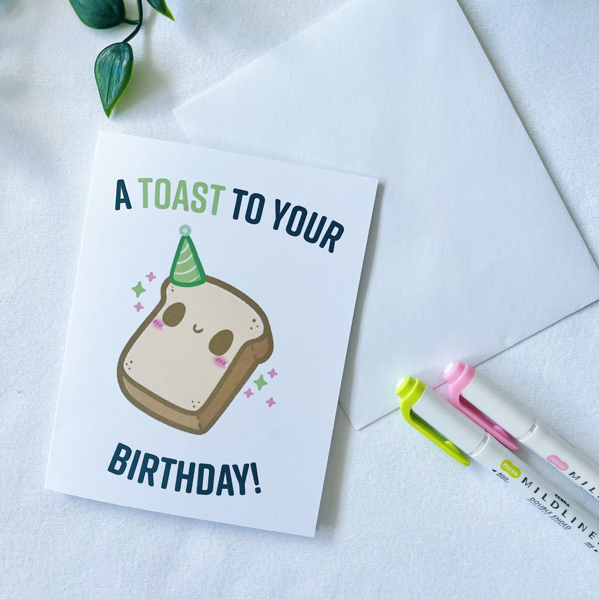 A white greeting card with a cute toast illustration and the text 'A TOAST TO YOUR BIRTHDAY!' is styled on a white surface with an envelope, green leaves, and two highlighters.