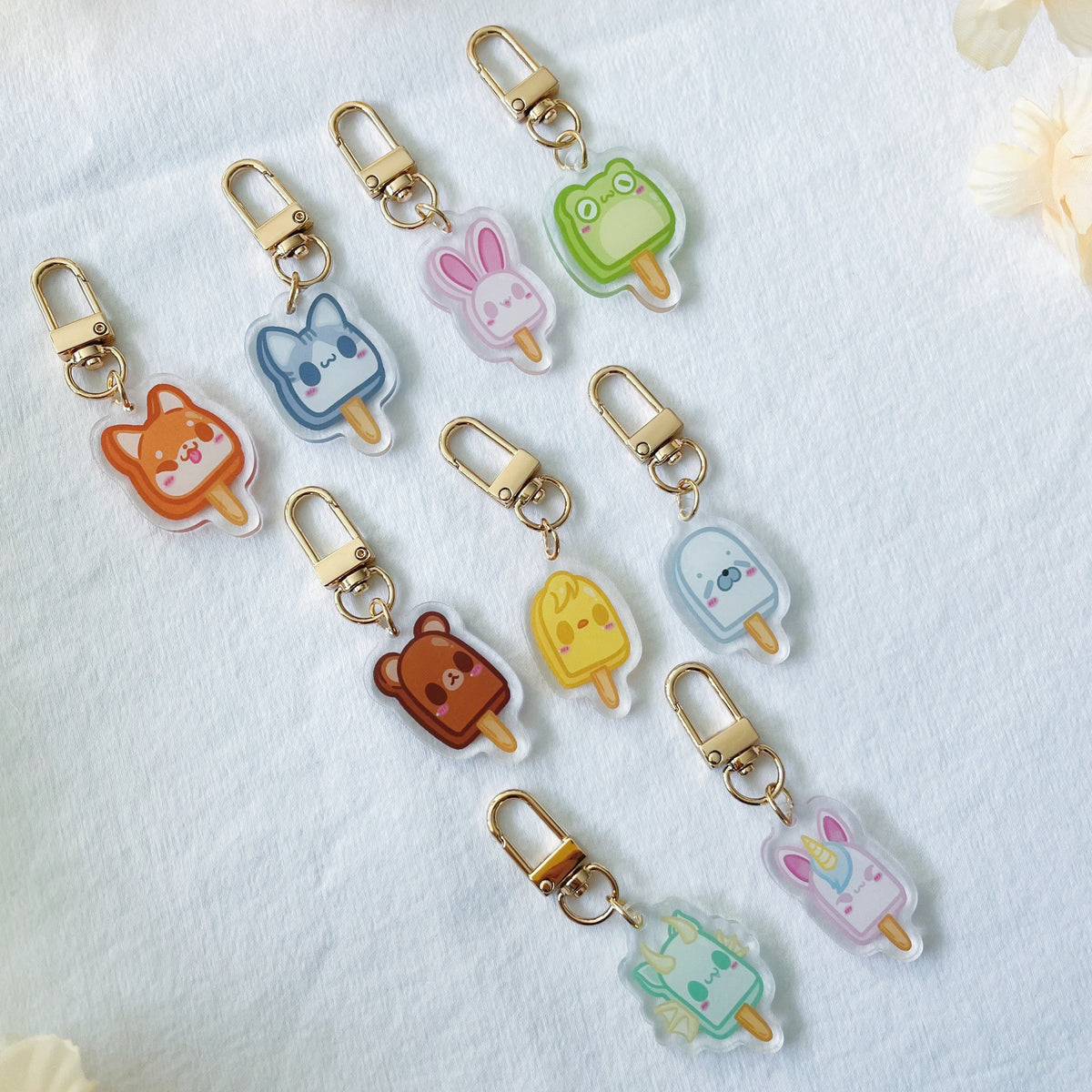 Six cute acrylic keychains shaped like animal popsicles (cat, bear, bunny, frog, duck, rabbit) in various colors, each with a gold-tone lobster clasp, arranged on a white textured fabric background with soft cream flowers in the corners.