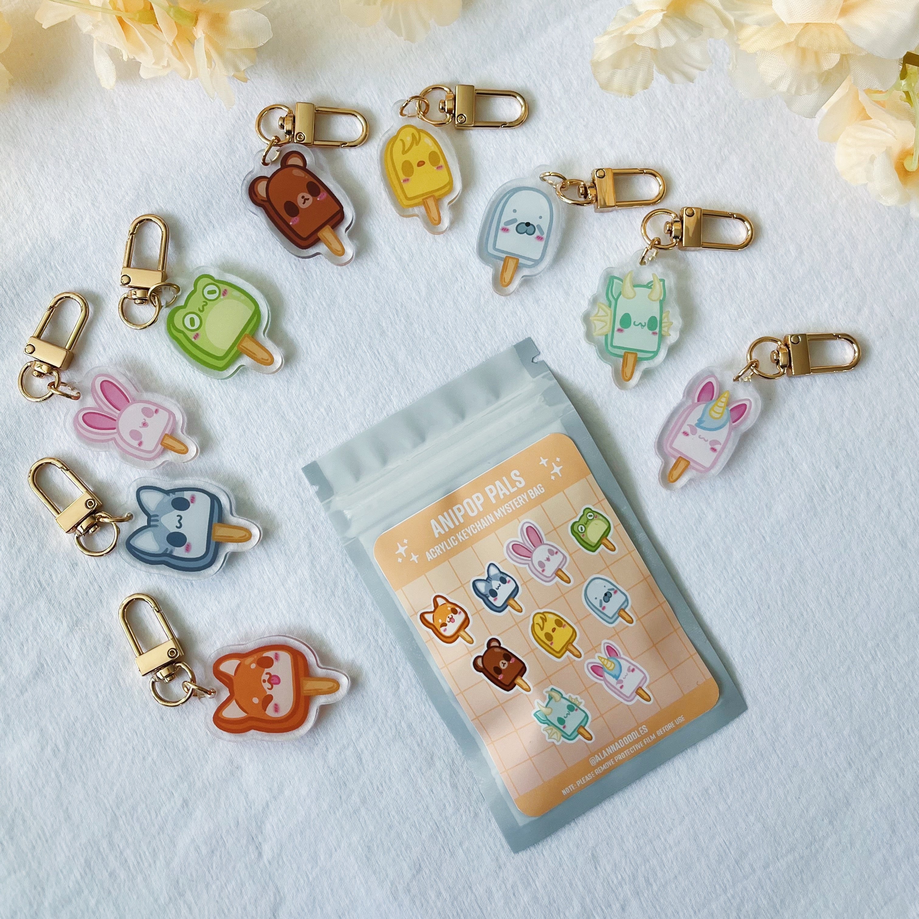 A set of cute, character-shaped acrylic keychains with gold-tone clasps, arranged around a small clear pouch showing the character sticker sheet. The keychains feature various characters like a frog, bunny, cat, and bear in pastel colors, set against a white textured background with soft yellow flowers.