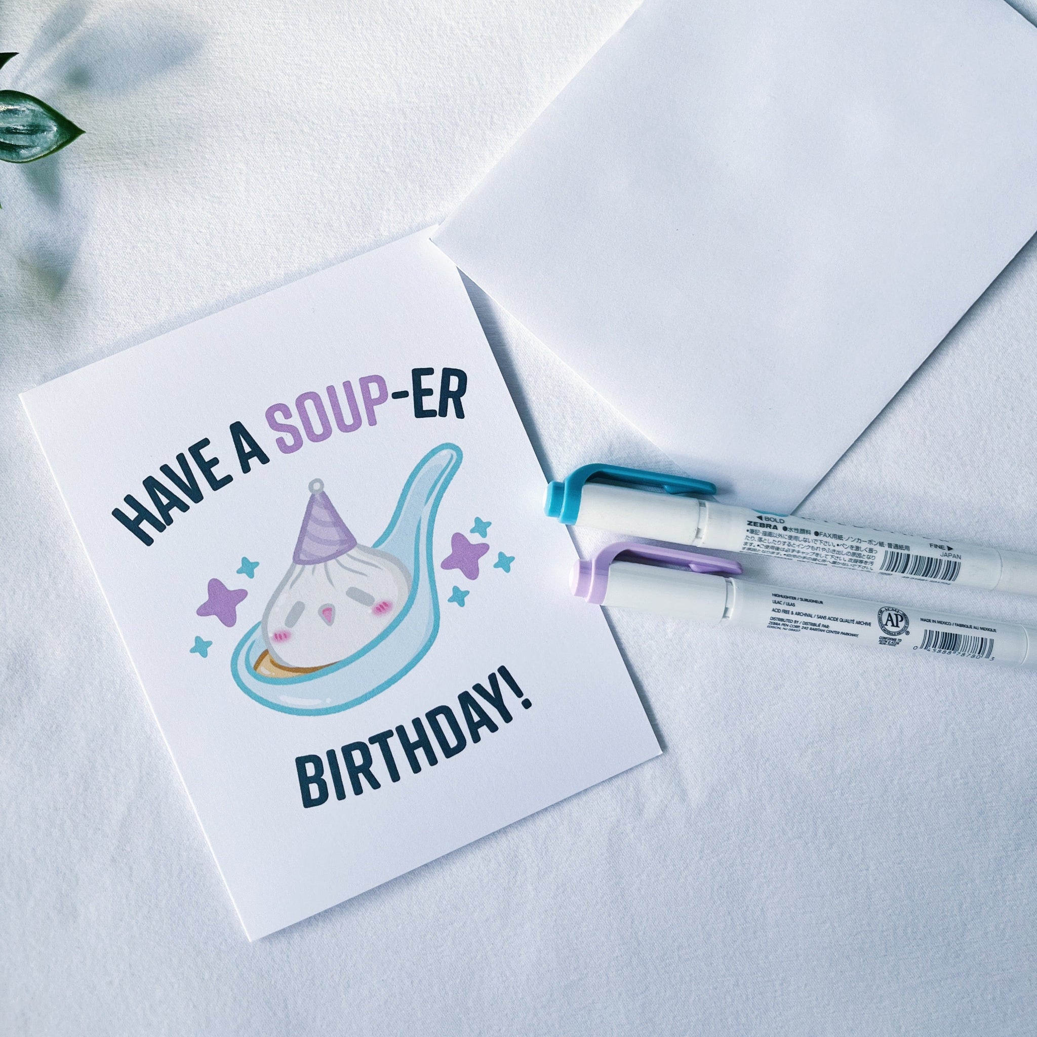 A square birthday greeting card with the text 'HAVE A SOUP-ER BIRTHDAY!' and a soup bowl illustration, shown on a white textured surface with an envelope and two markers nearby.