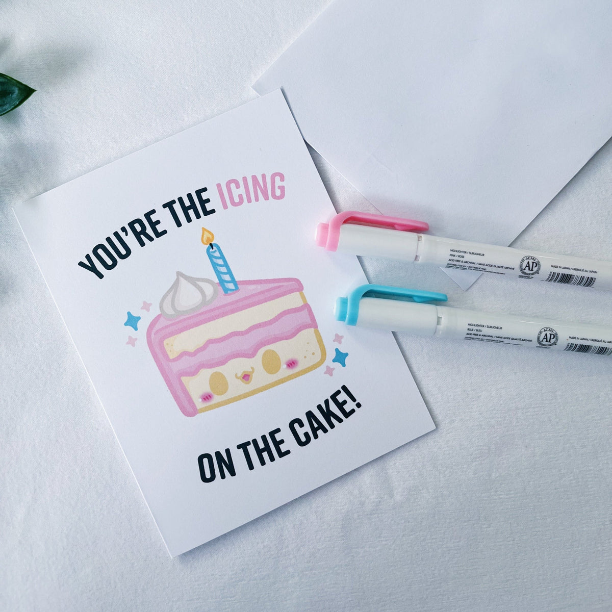 A square greeting card with a cute cake illustration and the text 'YOU'RE THE ICING ON THE CAKE!' is styled on a white textured surface with two markers and an envelope.